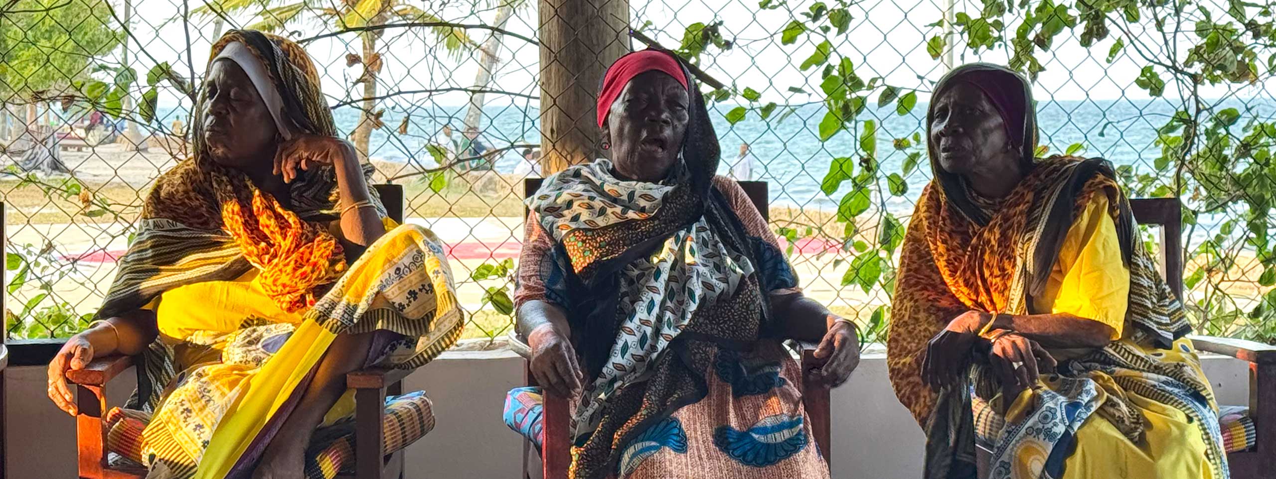 Drei ältere Frauen in bunten Kleidern sitzen am Meer.