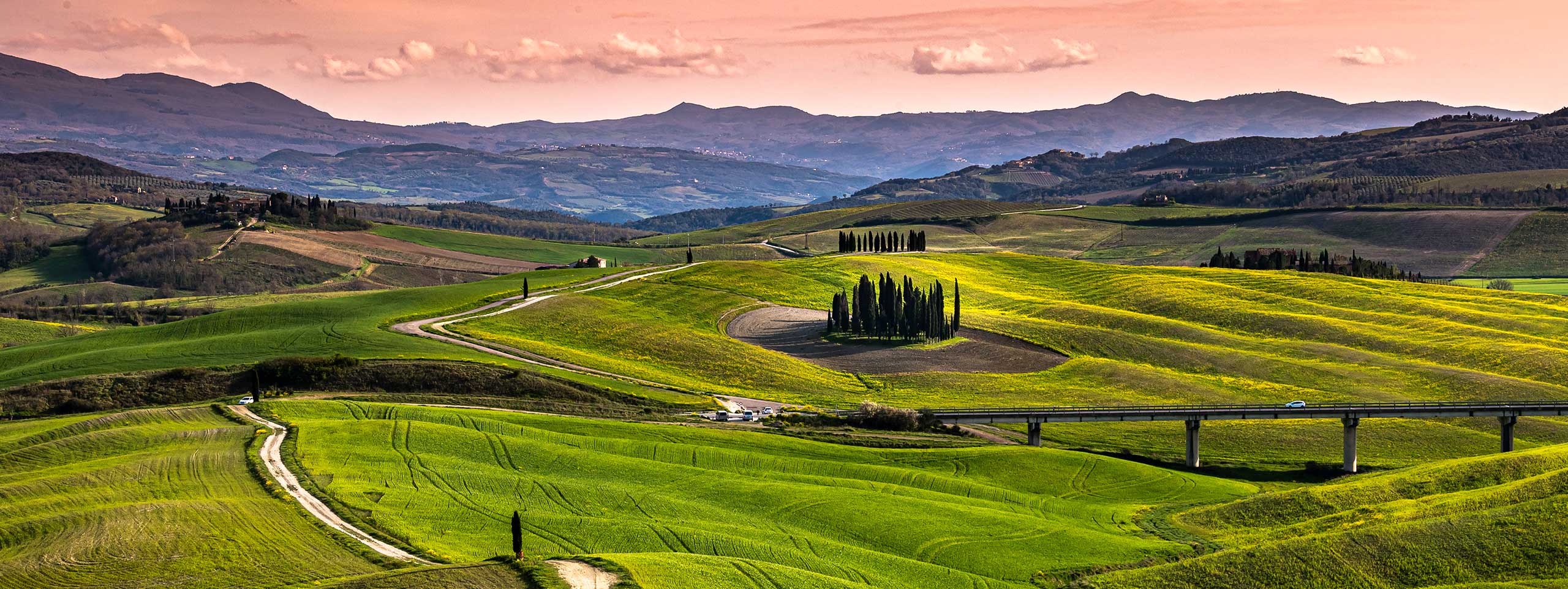 Grüne Hügellandschaft mit Zypressen, Straßen und Brücke.