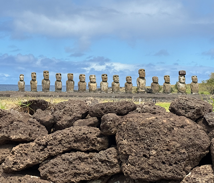 15 Moai-Statuen am Meer, dunkle Felsen davor.